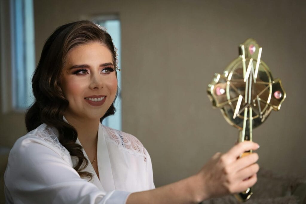 A woman smiles while holding a decorative hand mirror indoors, creating a warm reflective moment.